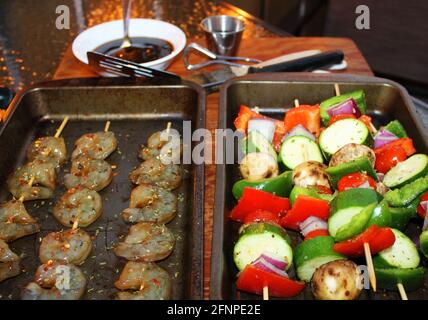 Nahaufnahme von Garnelen und Gemüsespiessen in Pfannen auf einem Schneidebrett, bevor sie gegrillt werden. Grillsoße und Geschirr im Hintergrund nicht im Fokus. Stockfoto