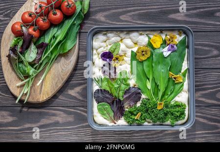 Sauerteig bereitet sich darauf vor, trendige Focaccia-Brote aus dem Garten auf dem Holztisch mit Bärlauch, essbaren Blumen und Olivenöl zu backen. Draufsicht. Stockfoto