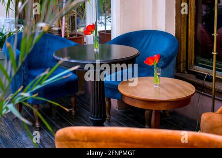 Terrasse eines Cafés im Freien mit weichen blauen Stühlen und Ein runder schwarzer Holztisch auf dem Holzboden von Das Fenster und Pflanzen mit Tulpen in Vasen a Stockfoto