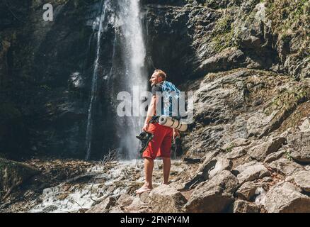 Mann mit Rucksack in aktiver Trekkingkleidung, die Trekking hält Stiefel in der Hand in der Nähe von Mountain River Wasserfall lächelnd und genießend The Nature.Trav Stockfoto