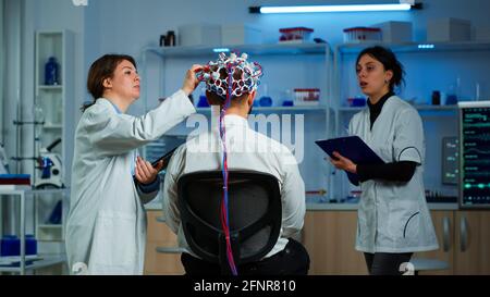 Ein Patient trägt ein leistungsfähiges eeg-Headset und scannt die elektrische Aktivität des Gehirns im neurologischen Forschungslabor, während ein medizinischer Forscher das Gehirn kontrolliert, das Nervensystem untersucht und die Krankenschwester Notizen macht Stockfoto