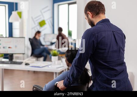Ein Geschäftsmann hilft seinem behinderten schwarzen Kollegen, der in das Firmenbüro eintritt und den Rollstuhl in die Nähe des Schreibtisches schiebt. Afrikanisch behinderte gelähmte Frau begrüßt Kollegen, die bei einem Finanzprojekt arbeiten. Stockfoto