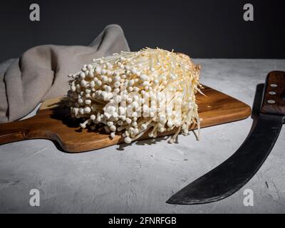 Frische goldene Nadelpilze oder Enoki-Pilze, auf Holzschneidebrett mit Vintage-Messer auf grauem Betongrund Stockfoto