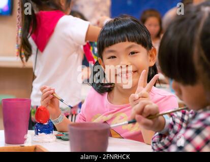 Portrait Asian Kind Mädchen in der Kunstwerkstatt, glücklich und lächelnd Gesicht, zeigen Geste zwei Finger, nettes Gesicht mit schwarzen Haaren und Knall. Verschwommener Klassenkamerad für Stockfoto