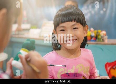 Portrait Asian Kind Mädchen in der Kunst-Werkstatt, glücklich und lächelnd Gesicht, schönes Lächeln, nettes Gesicht mit schwarzen Haaren, und Knall. Verschwommener Klassenkamerad im Vordergrund. Stockfoto