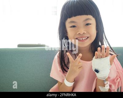 Asiatische Kind Mädchen sitzt auf Krankenhausbett, zeigt zwei Finger. Positives Bild des Patienten im Krankenhaus. Stockfoto