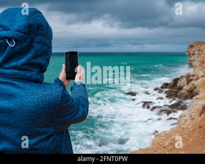 Eine Frau fotografiert das Meer auf dem Smartphone, eine Frau reist in der kalten Jahreszeit entlang der Küste Stockfoto