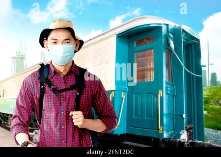 Asiatischer Mann in der Gesichtsmaske mit Hut und Rucksack auf dem Bahnhof. Reisen in der neuen Normalität Stockfoto
