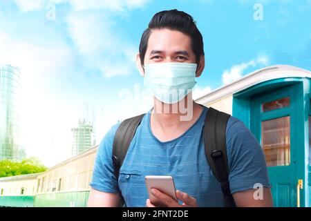 Asiatischer Mann in der Gesichtsmaske mit einem Rucksack, der das Smartphone am Bahnhof hält. Reisen in der neuen Normalität Stockfoto