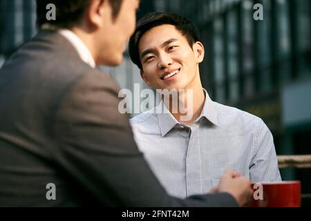 Zwei asiatische Geschäftsleute, die bei einem Kaffee im Freien über Geschäfte diskutieren Kaufen Stockfoto