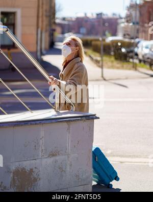 Modische ältere Frau, die in einem Atemschutzgerät oder einer Gesichtsmaske auf einer sonnigen Straße mit blauem Koffer steht und nach oben schaut. Ältere Dame verirrt sich während der Reise während des covid19-Ausbruchs in der Stadt Stockfoto