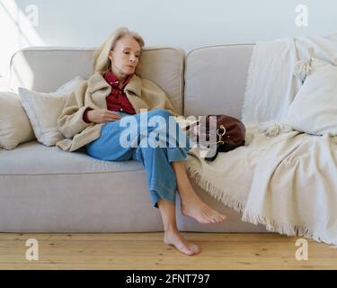 Müde reife 60s Frau in Freizeitkleidung ruhen nach der Arbeit auf der Couch zu Hause, beiseite schauen und denken oder darüber nachdenken. Nachdenkliche ältere Dame, die nach einem harten Arbeitstag alleine auf dem Trainer sitzt Stockfoto