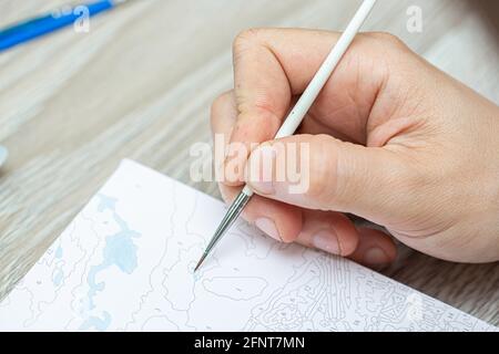 Eine mans Hand hält einen Pinsel und zeichnet ein Bild durch Zahlen. Zeichnen auf Leinwand. Nummerierte Farbdosen. Kreatives Hobby. Malen für Anfänger.Nahaufnahme, sele Stockfoto