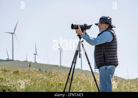 Älterer Mann mit neuer Technologie. Kamera auf Stativ, lässig gekleideter Mann. Windturbinen im Hintergrund. Abruzzen, Italien, Europa Stockfoto