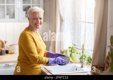 Fröhliche ältere Dame, die in der Küche mit Gummihandschuhen Geschirr abspülte Stockfoto