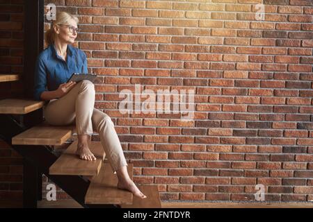 Entspannte, reife, blonde kaukasische Frau, die eine Brille trägt, die wegschaut und einen Tablet-pc benutzt, während sie bequem auf einer Holztreppe vor der Ziegelwand sitzt Stockfoto