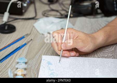 Eine mans Hand hält einen Pinsel und zeichnet ein Bild durch Zahlen. Zeichnen auf Leinwand. Nummerierte Farbdosen. Kreatives Hobby. Malen für Anfänger.Nahaufnahme, sele Stockfoto