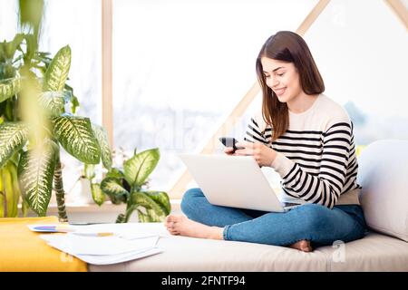 Aufnahme einer jungen Frau, die ihren Laptop und SMS verwendet, während sie von zu Hause aus arbeitet. Heimbüro. Stockfoto