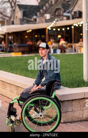 Ein junger Mann in einer behinderten Person sitzt auf einer Bank neben einem Rollstuhl Stockfoto