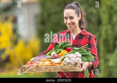 Schöne dunkelhaarige Bauernfrau hält den Holzkorb voll mit frischem, gesundem Gemüse aus dem Garten. Stockfoto