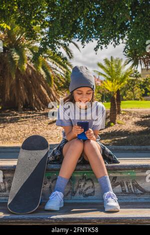 Das Mädchen aus der Vorpubertät, kaukasisch, sitzt auf einem Hindernis in einem Skatepark, mit den Füßen auf ihrem Brett, während sie mit ihrem Mo chattet oder im Internet surft Stockfoto