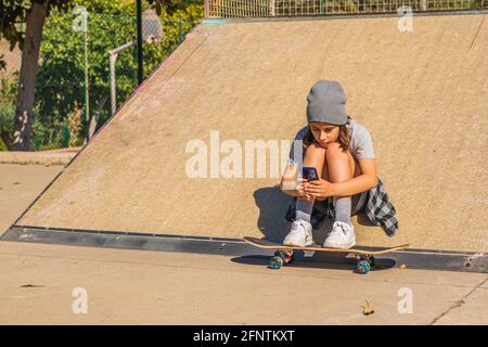 Kaukasisches Mädchen, das auf einer Skateboard-Rampe sitzt und ihr Brett neben sich hat, während es auf ihr Mobiltelefon schaut Stockfoto