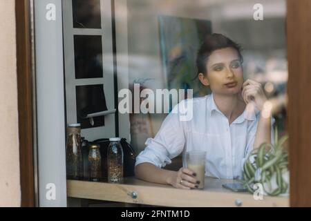 Ein schönes junges Mädchen sitzt in einem Café hinter einem Glas mit einer Tasse Kaffee und schaut nachdenklich aus dem Fenster. Foto durch Glas mit Straßenrefle Stockfoto