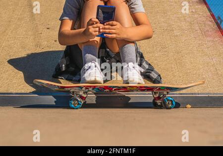 Unkenntlich kaukasisches Mädchen, das auf einer Skateboard-Rampe mit ihrem Brett neben sich sitzt, während es auf ihr Mobiltelefon schaut Stockfoto