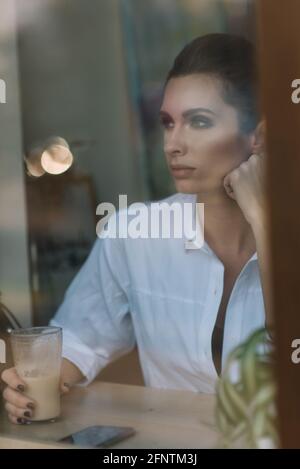 Ein schönes junges Mädchen sitzt in einem Café hinter einem Glas mit einer Tasse Kaffee und schaut nachdenklich aus dem Fenster. Foto durch Glas mit Straßenrefle Stockfoto