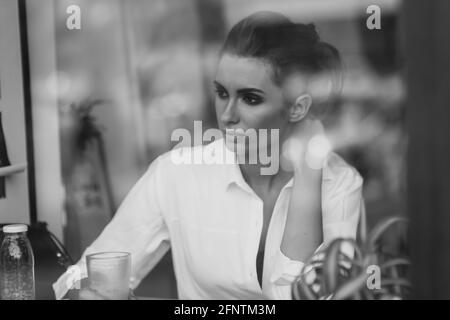 Ein schönes junges Mädchen sitzt in einem Café hinter einem Glas mit einer Tasse Kaffee und schaut nachdenklich aus dem Fenster. Schwarzweiß-Kunstfoto durch gla Stockfoto