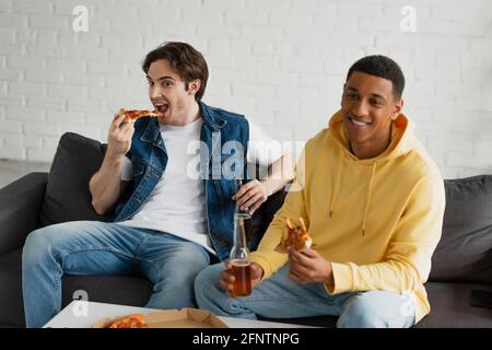 Interrassische Freunde genießen Pizza und Bier, während sie auf der Couch sitzen In modernem Loft Stockfoto