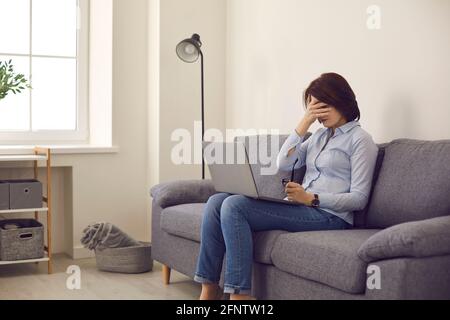 Die Frau, die auf dem Sofa sitzt und den Laptop dabei hat, hört auf zu arbeiten und deckt sich ab Gesicht fühlt sich müde und unglücklich Stockfoto