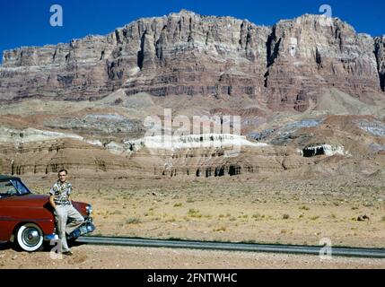 Junger Mann, der sich in den 1950er Jahren auf einen roten Plymouth Cabriolet mit weißen Felgen aus dem Jahr 1952 stützte. Das Auto steht am Straßenrand neben den Painted Desert Cliffs, Arizona, USA in den 1950er Jahren Stockfoto