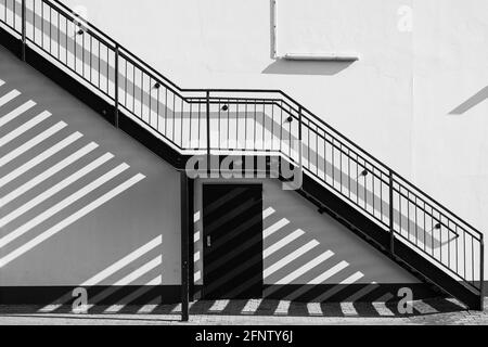 Eine Außentreppe verläuft schräg entlang der Hauswand und wirft Schatten auf die Wand und den gepflasterten Boden - in Schwarz und Weiß. Stockfoto