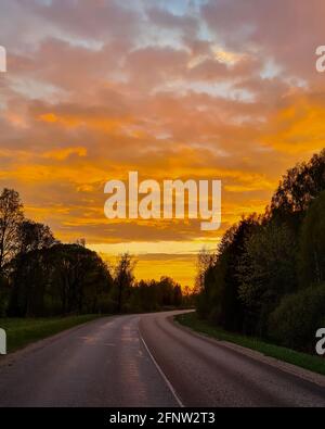 Asphaltstraße in der Landschaft bei orangefarbenem Sonnenuntergang. Neben grünen, frühlingsblühenden Bäumen. Stockfoto