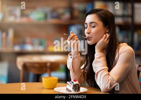 Glückliche arabische Dame, die Dessert und Kaffee mit geschlossenen Augen genießt, im Café sitzt und bei der Arbeit Pause macht Stockfoto