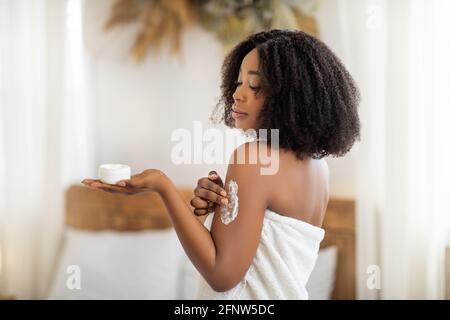Schönheitsbehandlung für gesunde Haut. Wunderschöne schwarze Frau im Badetuch Anwendung Körpercreme oder Butter auf der Schulter zu Hause Stockfoto