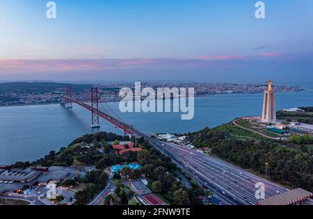 Luftaufnahme einer mehrspurigen Straße, die in der Dämmerung auf einer Hängebrücke über den Fluss Tejo führt, Lissabon, Portugal. Stockfoto