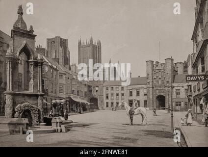 Eine Ansicht des Market Place aus dem späten 19. Jahrhundert in Wells, einer Stadt im Mendip-Viertel von Somerset, England. Der Market Place, ein Ort eines der alten Brunnen der Stadt (man beachte das Wasser, das in der Schlucht neben dem Bürgersteig fließt); das Bishop's Eye Torhaus (zum Bishop's Palace) befindet sich am anderen Ende; zwei der Türme der Wells Cathedral sind hinter den Gebäuden zu sehen. Stockfoto