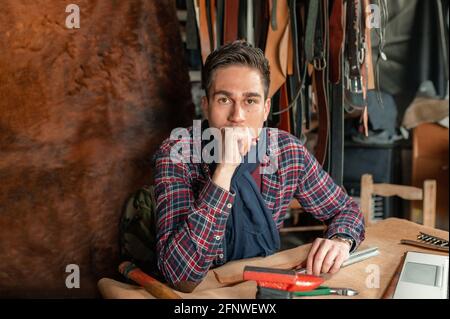 Porträt eines selbstbewussten jungen Mannes, der am Schreibtisch in seinem Labor sitzt und die Kamera anschaut. Arbeiten Sie mit echtem Leder, Kunsthandwerk und Hobbys zu Hause c Stockfoto