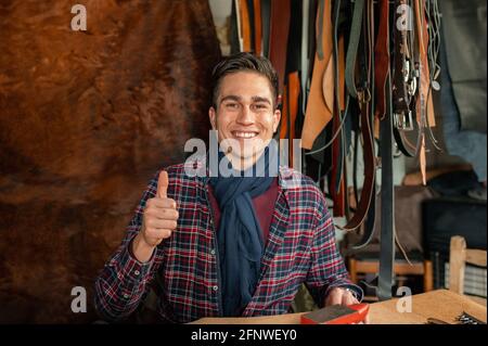Porträt eines selbstbewussten lächelnden jungen Mannes, der am Schreibtisch in seinem Labor sitzt und die Kamera anschaut und den Daumen nach oben zeigt. Arbeiten mit echtem Leder, Stockfoto