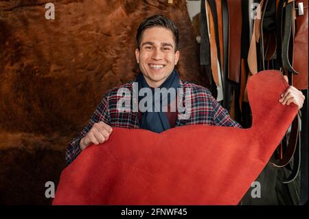 Lächelnder junger Mann, der ein Stück rotes Echtleder hält und die Kamera in seiner Werkstatt ansieht. Stockfoto