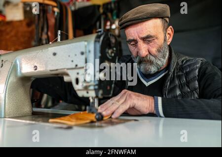 Älterer alter Hipster bärtiger Mann mit flacher Mütze, der mit Leder und Nähmaschine arbeitet. Stockfoto