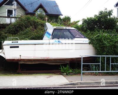 Russland, Sotschi 16.04.2021. Ein altes Motorboot steht auf der Straße auf einem Stand in der Nähe der Büsche und des Wohnhauses Stockfoto