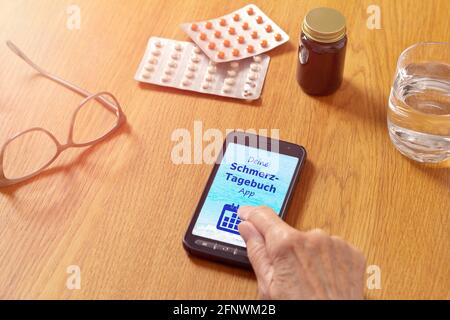 Medizin auf einem Tisch und eine Hand berühren ein Smartphone mit einer deutschen mobilen Anwendung, Text: Schmerztagebuch App. Übersetzung: Ihre Schmerztagebuch App. Stockfoto