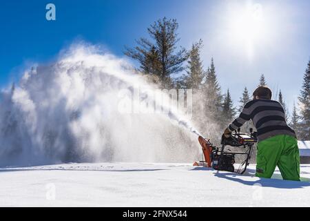 Weitwinkelansicht von hinten eines Mannes, der einen Schneepflug im knietiefen Neuschnee manövriert, wobei ein hoher Jet wie ein Schneesturm aus der Maschine austritt Stockfoto