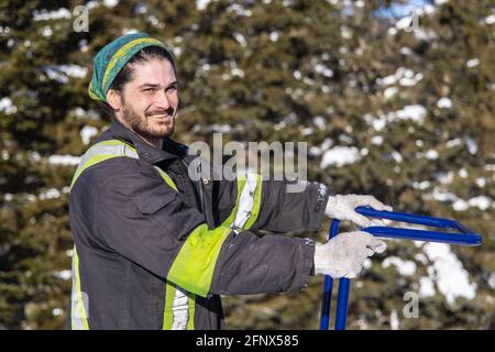 Porträt eines lächelnden jungen Mannes, der mit seinen Händen auf dem Griff einer blauen Handschaufel posiert, während er Neuschnee aus einem Landhaus entfernt. Stockfoto