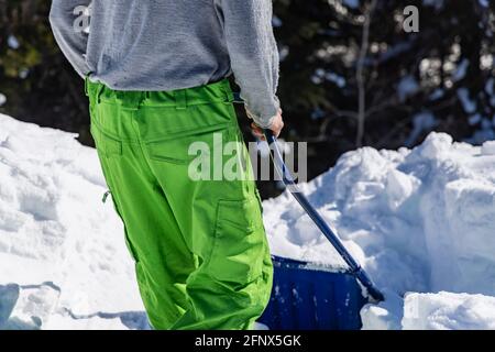 Blick von hinten auf die Hände und Beine eines jungen Mannes, der mit einer blauen Handschaufel frischen Schnee vom Dach eines Landhauses entfernt. Stockfoto