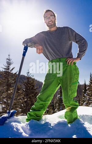 Porträt eines lächelnden Mannes, der sich auf den Griff einer blauen Schneeschaufel lehnt, während er während einer Schneeräumung in einem Landhaus eine Pause einnimmt. Stockfoto