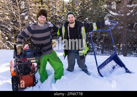 Portrait von zwei jungen Männern, die mit ihren Schneebesen, einem leuchtend orangenen mechanischen Schneepflug und einer blauen Handschaufel für die Kamera posieren. Stockfoto